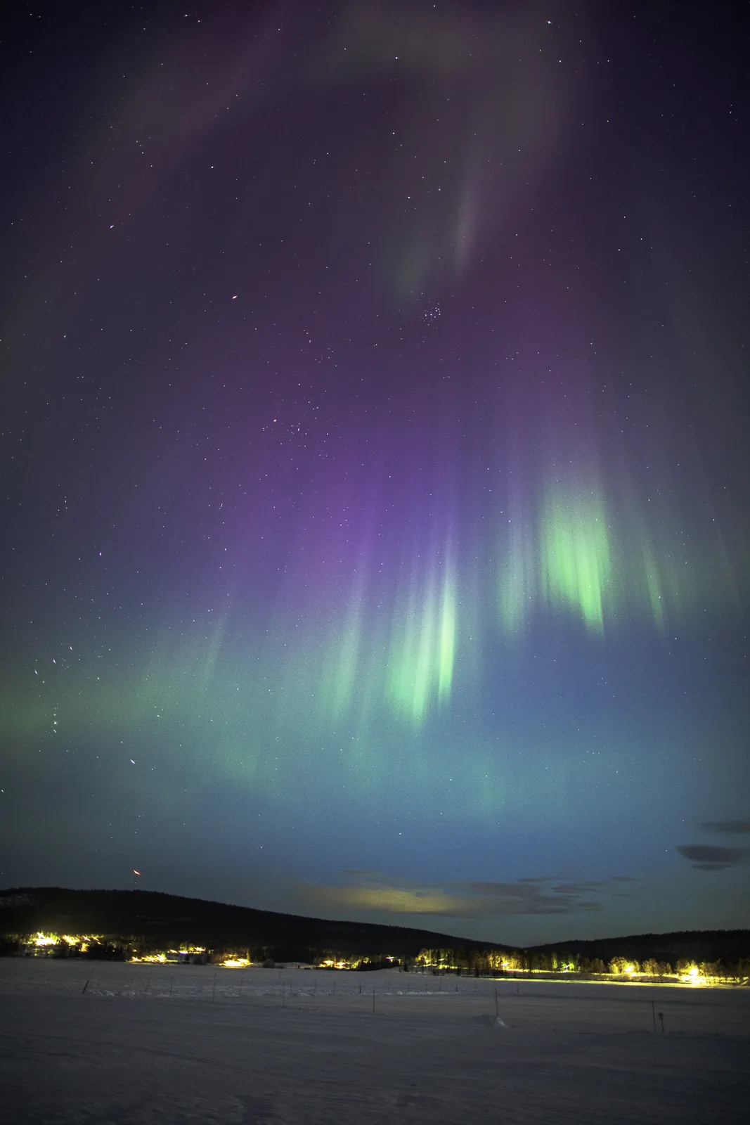Purple and green aurora over a frozen lake with village lights near Kiruna