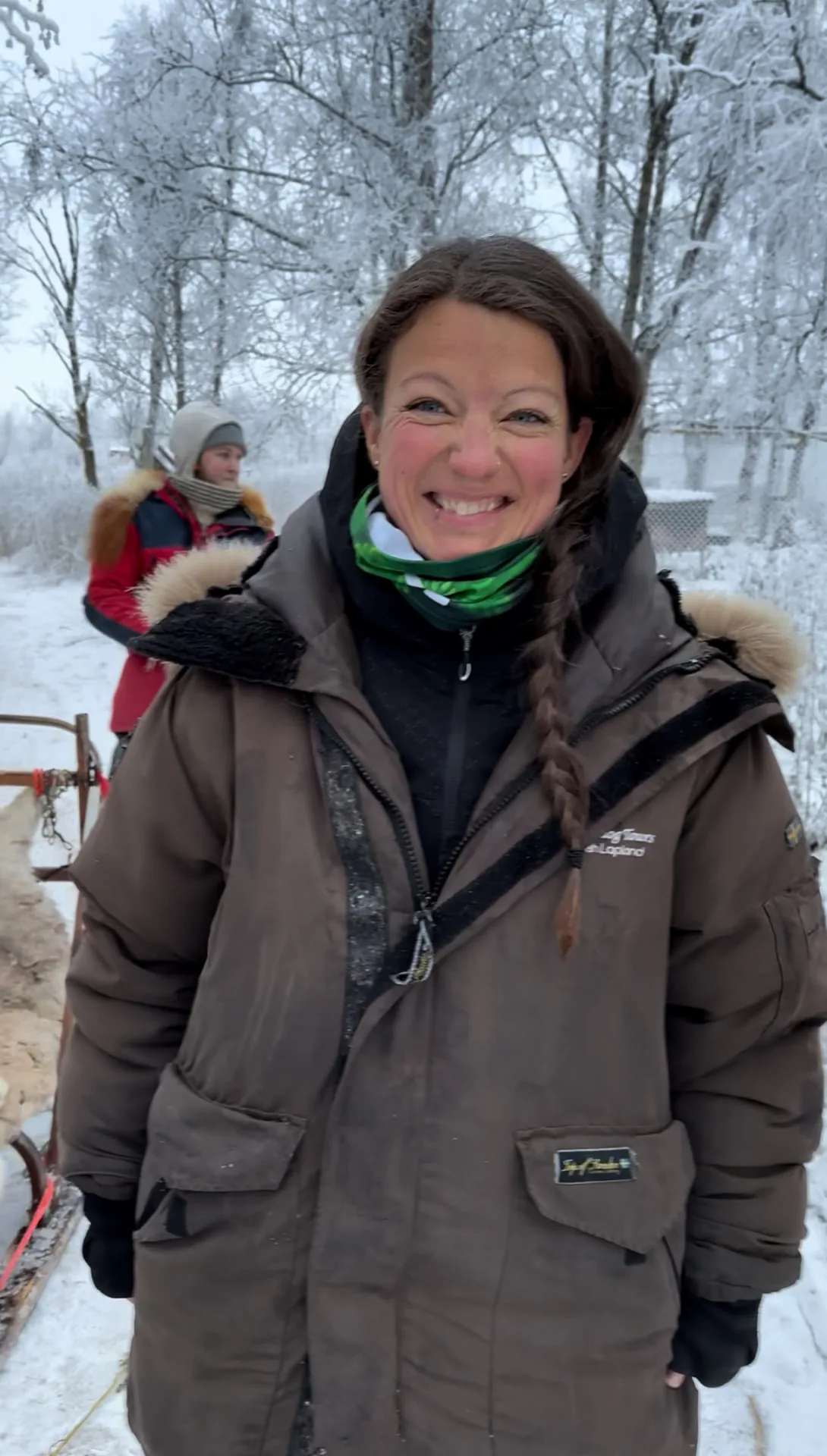 Christina Bihl smiling in a brown winter parka beside a dog sled on a frosted forest trail