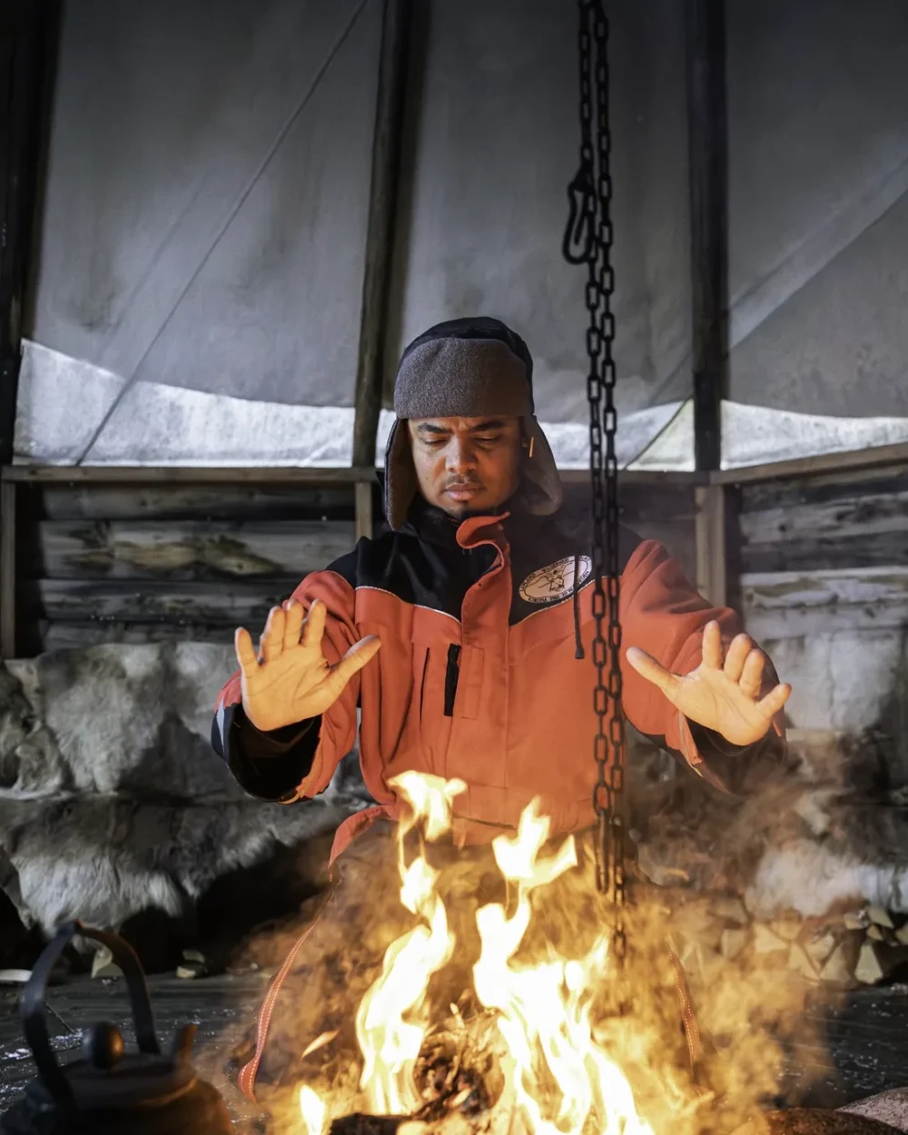 Justin Perez, a guest from the United States, warming his hands over the open fire inside the lavvu, wearing the winter clothing set we lend out
