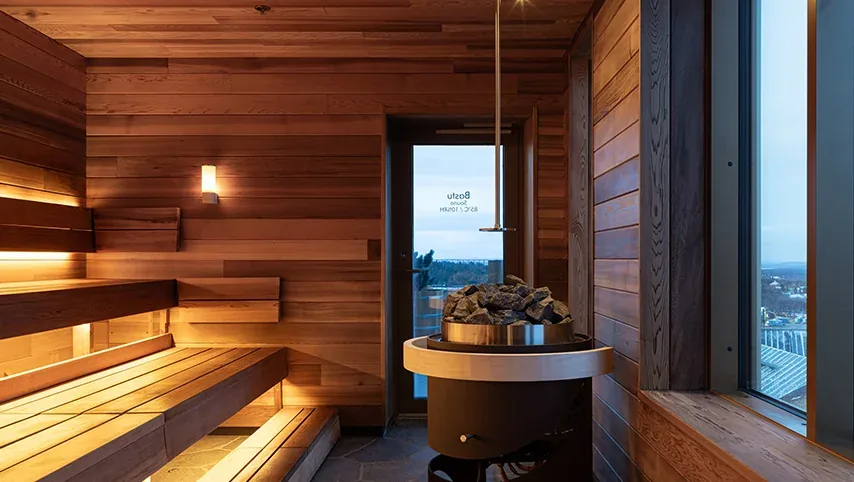 Cedar-lined sauna at Vana Spa with tiered benches, a stone-topped heater and a window over Kiruna at dusk